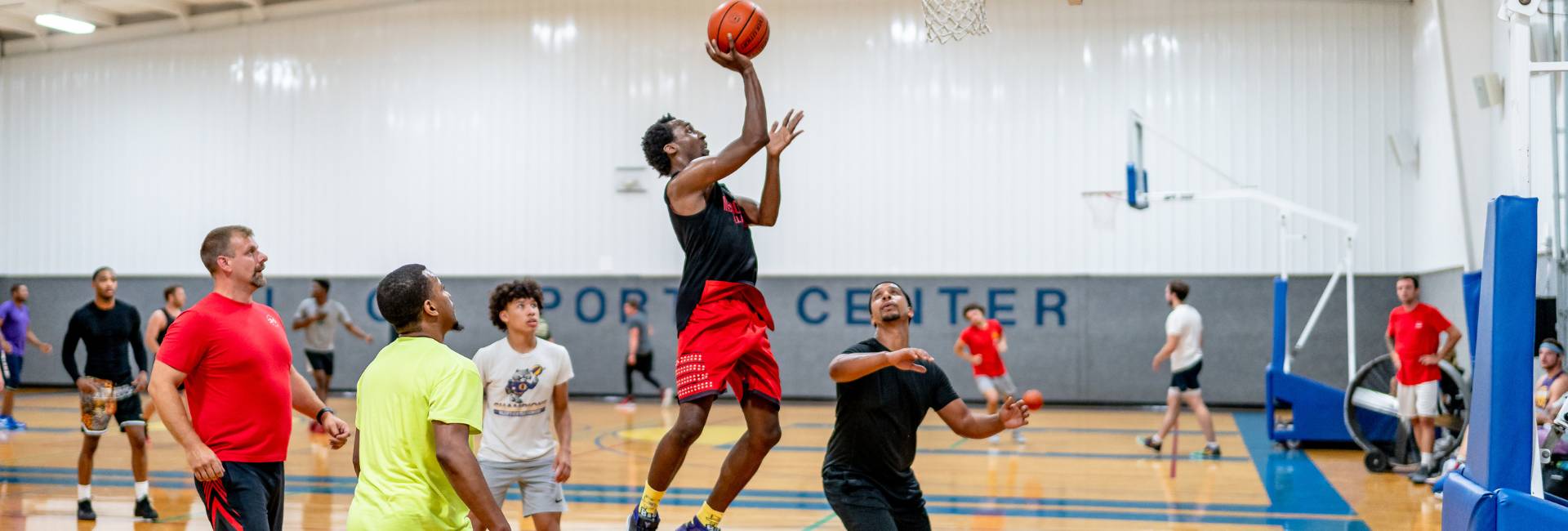 A basketball player in a red and black uniform jumps toward the hoop for a layup while others watch during an indoor game.